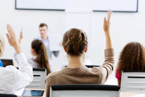 A group of men an women sit in a classroom. One woman has her hand raised to ask a question.